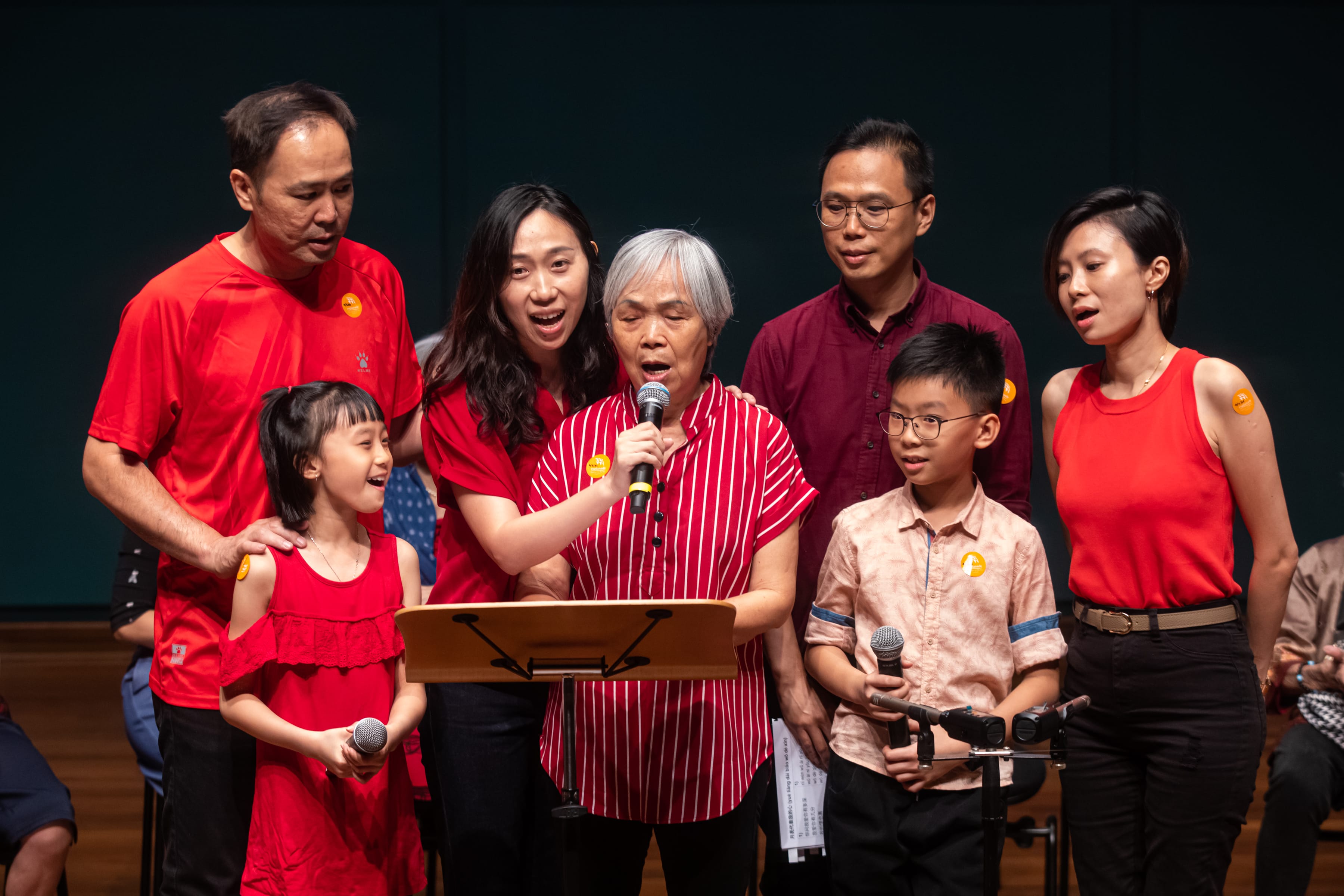 A senior stands in the centre singing surrounded by a group of people